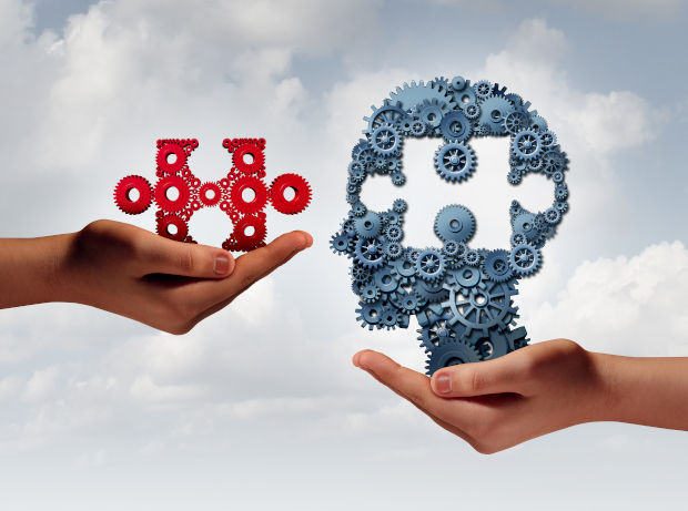 Two hands hold gear formations against a cloudy sky. The left hand holds red puzzle-shaped gears, the right holds a blue gear head shape. Symbolizes synergy.