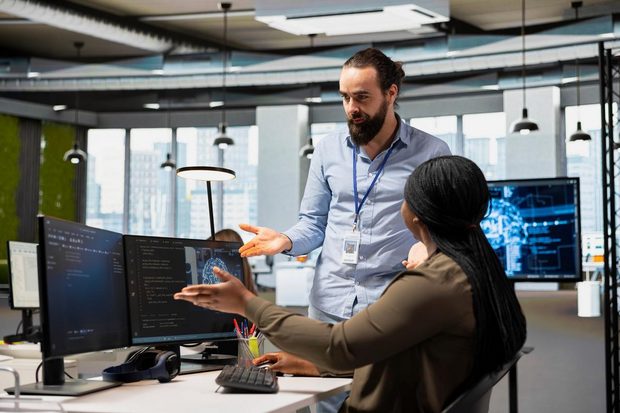 Two colleagues reviewing code and AI dashboards together at a shared desk.
