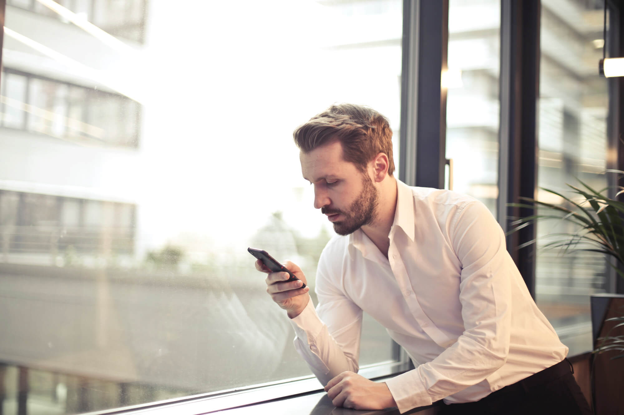 Blond-haired business man looking at mobile phone while leaning against a window