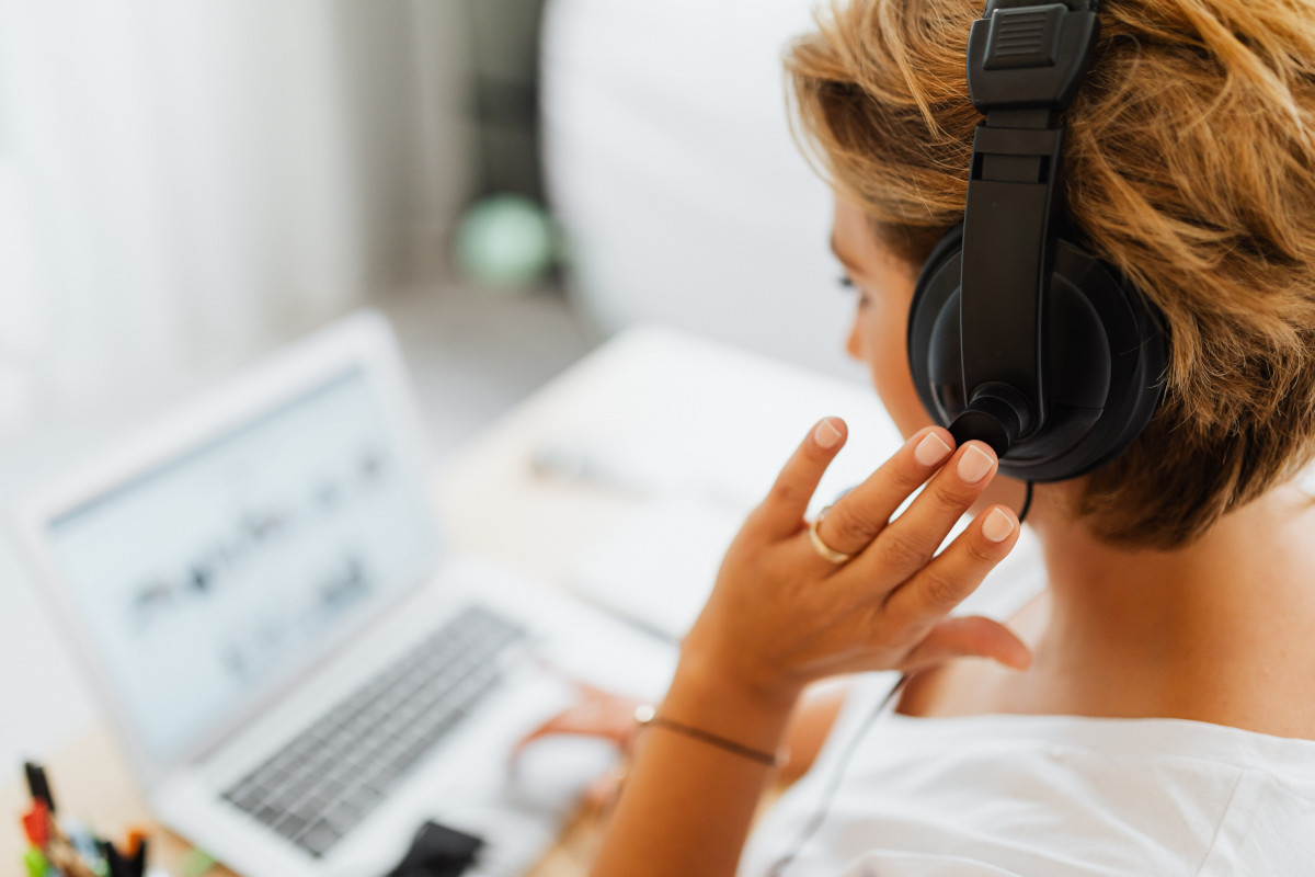 A woman in white shirt using black headphones while working on a laptop.