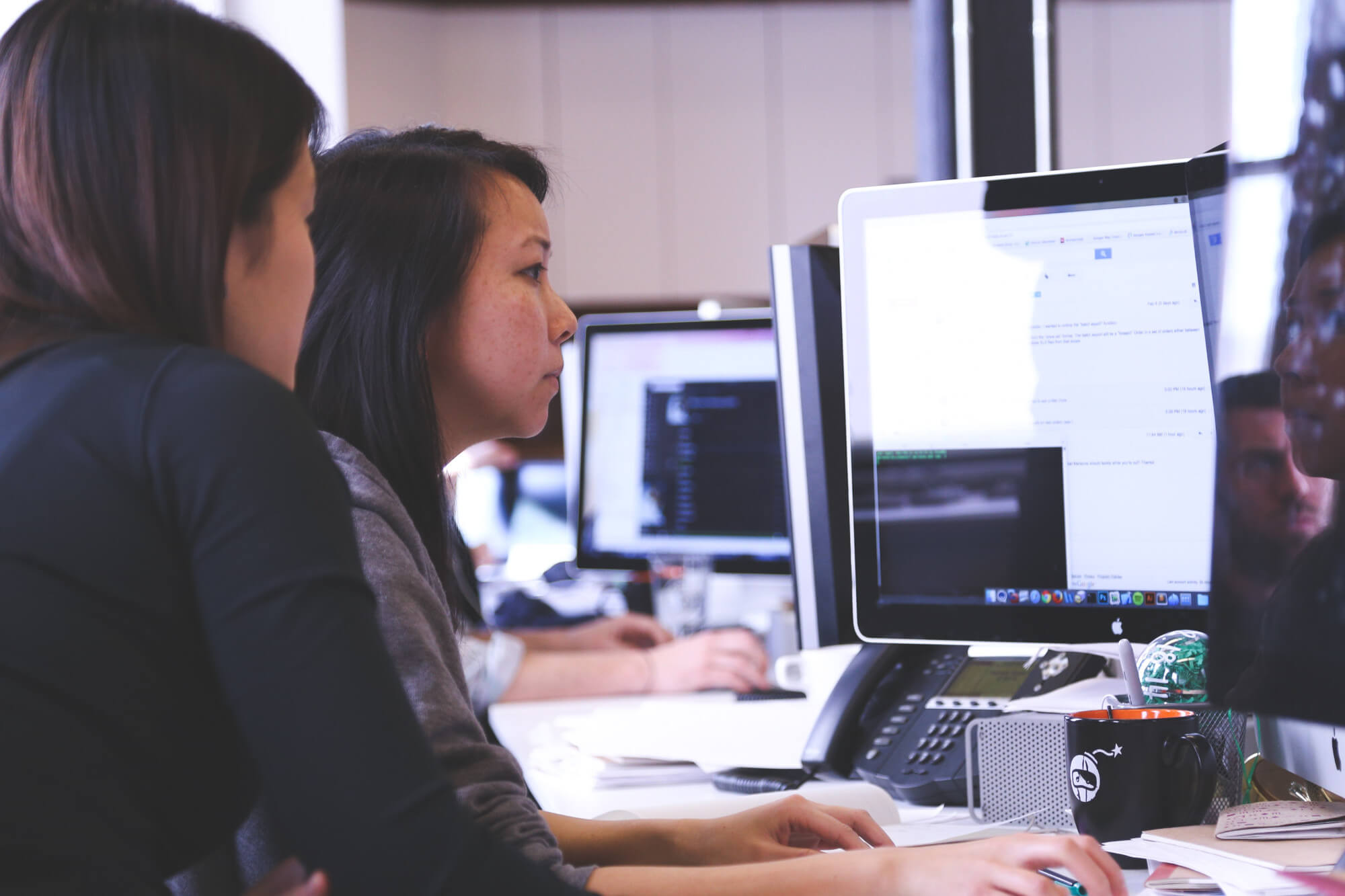 Two women working together in an office, in front of an Apple monitor