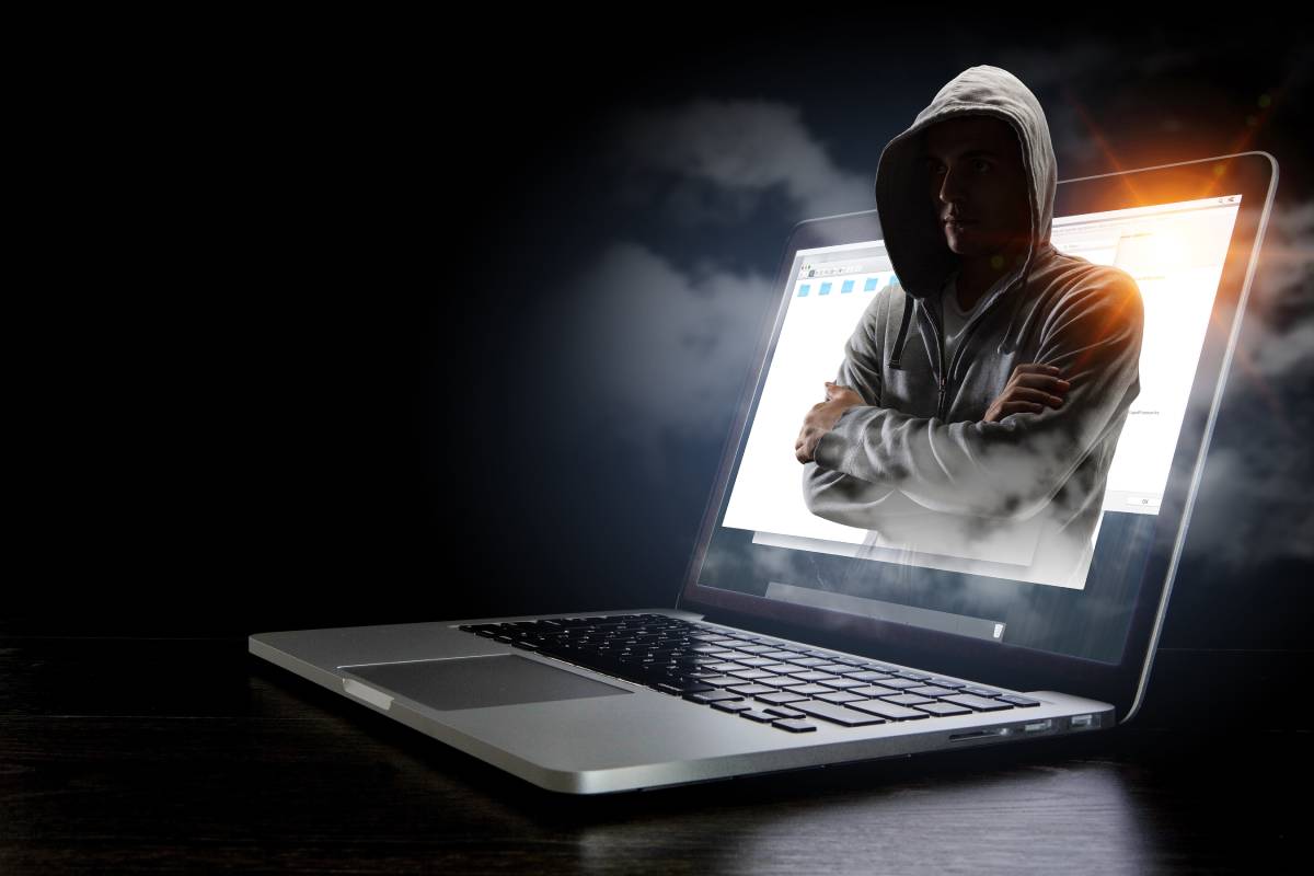 A hooded figure representing a hacker is coming out of a laptop screen