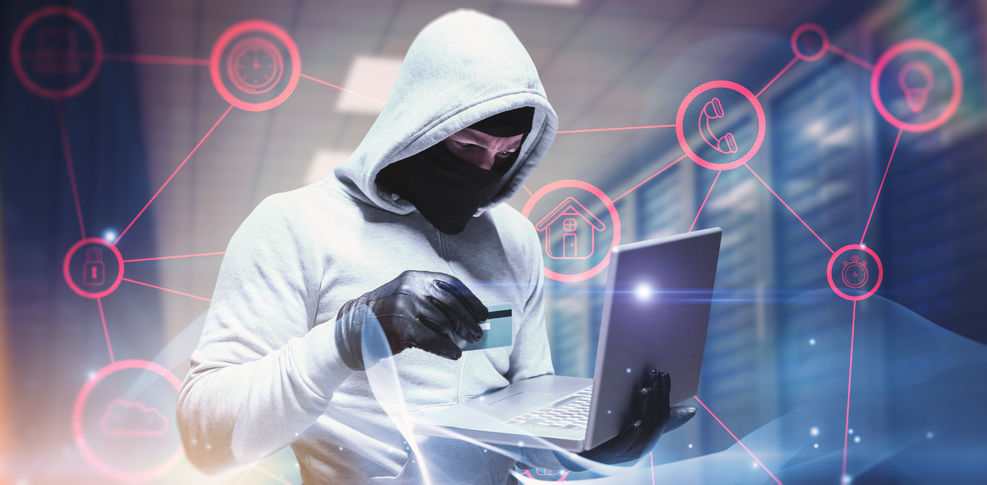 A hacker in a hoodie holding a credit card in one hand and a laptop in another while standing in a server room