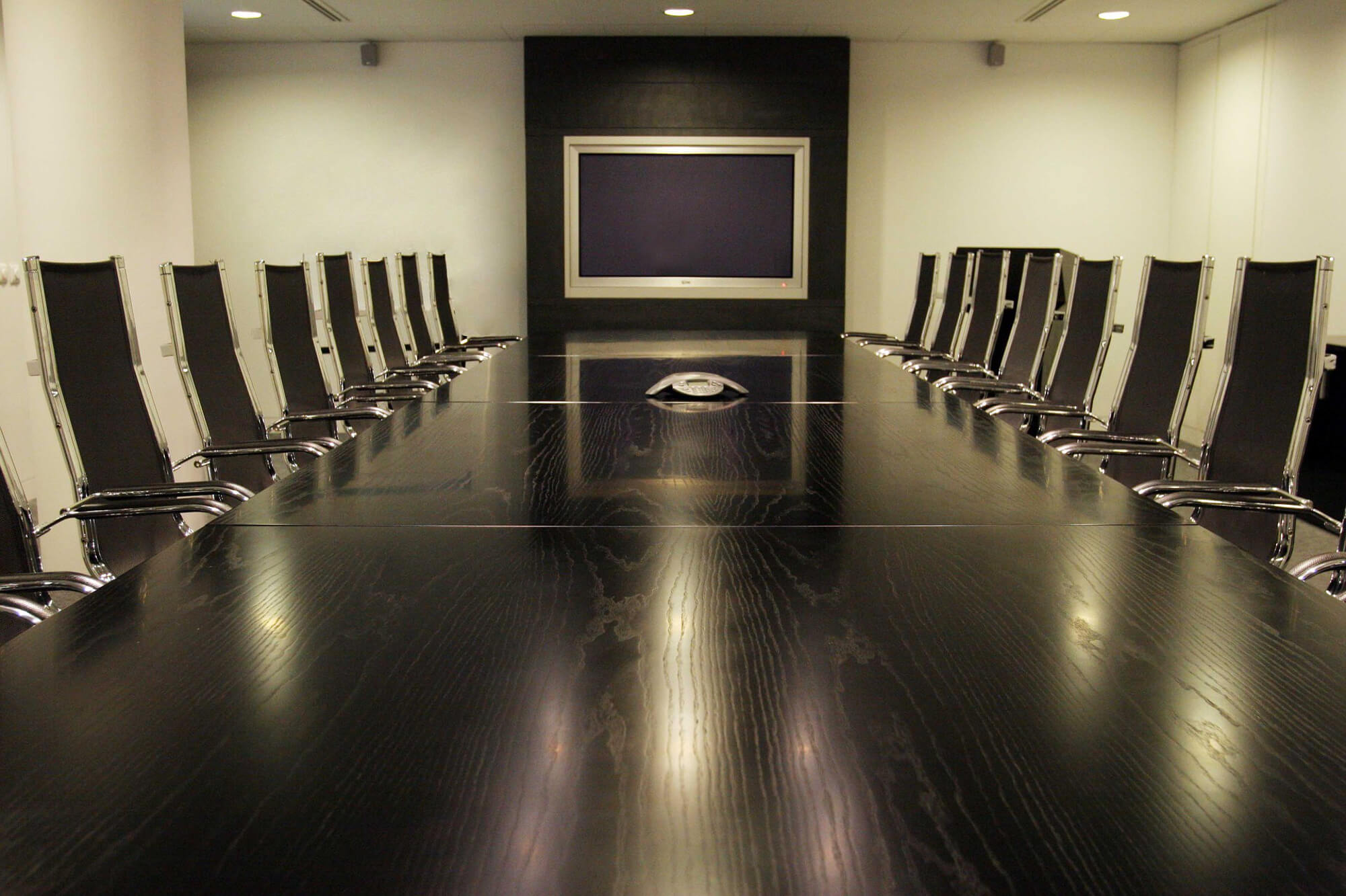 Conference room with dark wood table, black and silver chairs, a speaker phone, and a tv display on the far wall