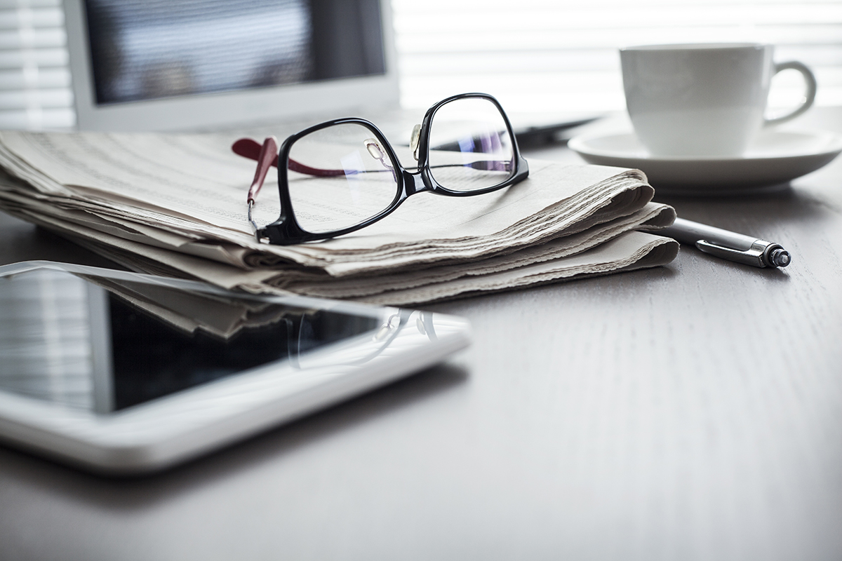 A tablet, a newspaper with glasses sitting on it, a pen, a coffee cup and a laptop sitting on a table.