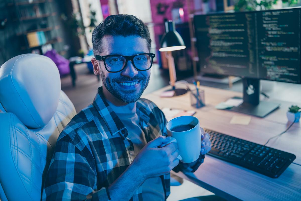 Person holding a coffee mug at a desk beside a monitor filled with code.