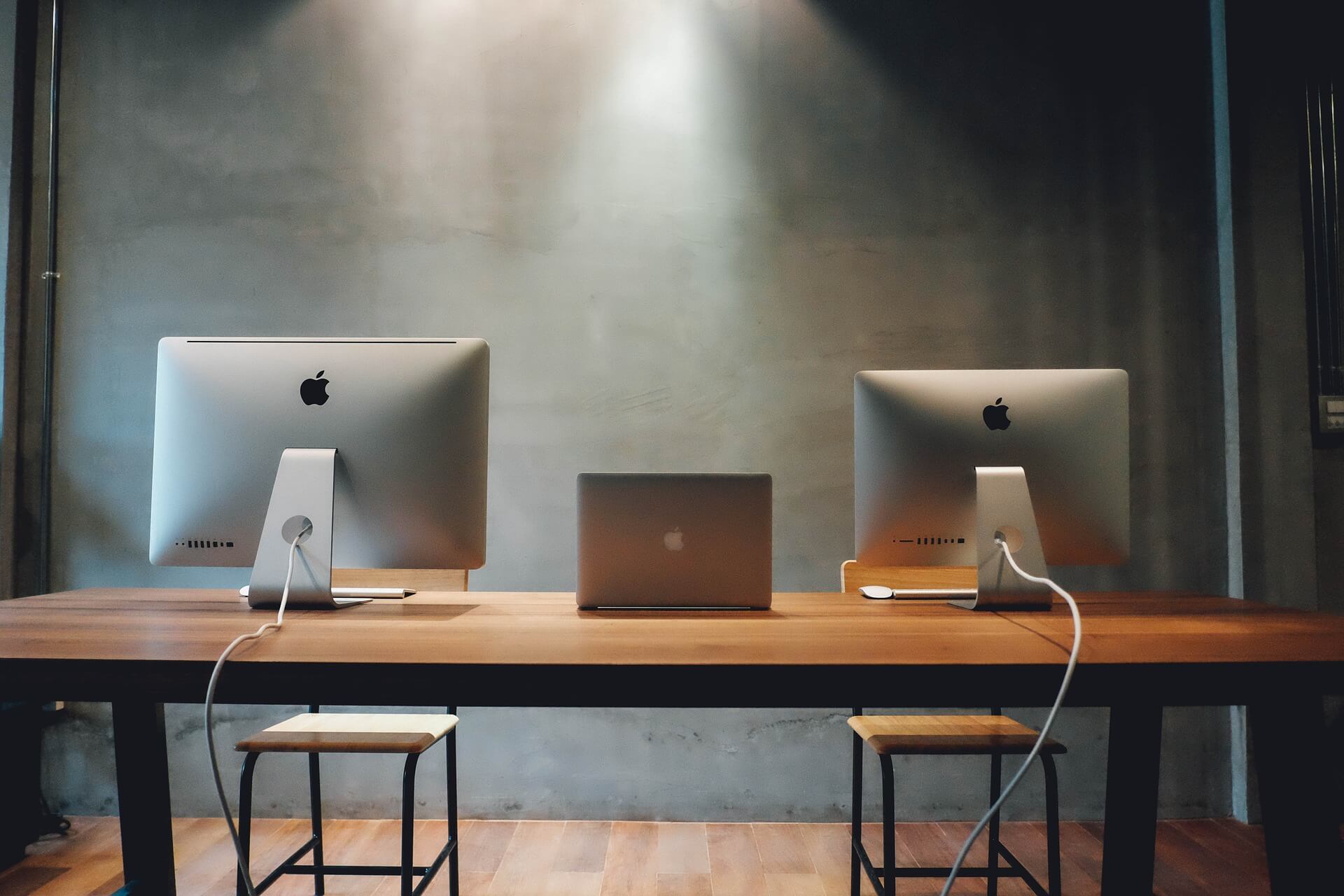 View of the backside of a wooden desk with 2 Apple computers and an Apple laptop, all in gray