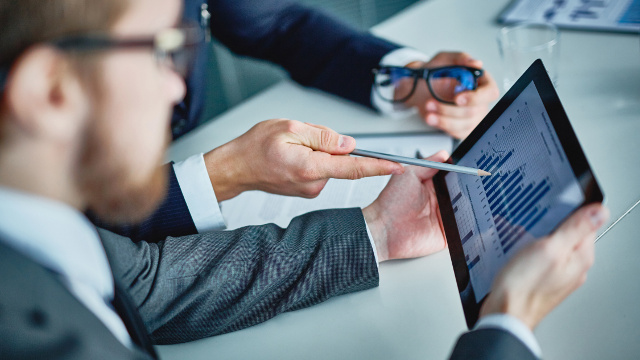 Businessman Gesturing with a Pencil while Bearded Man Blurred in Foreground Holds Tablet