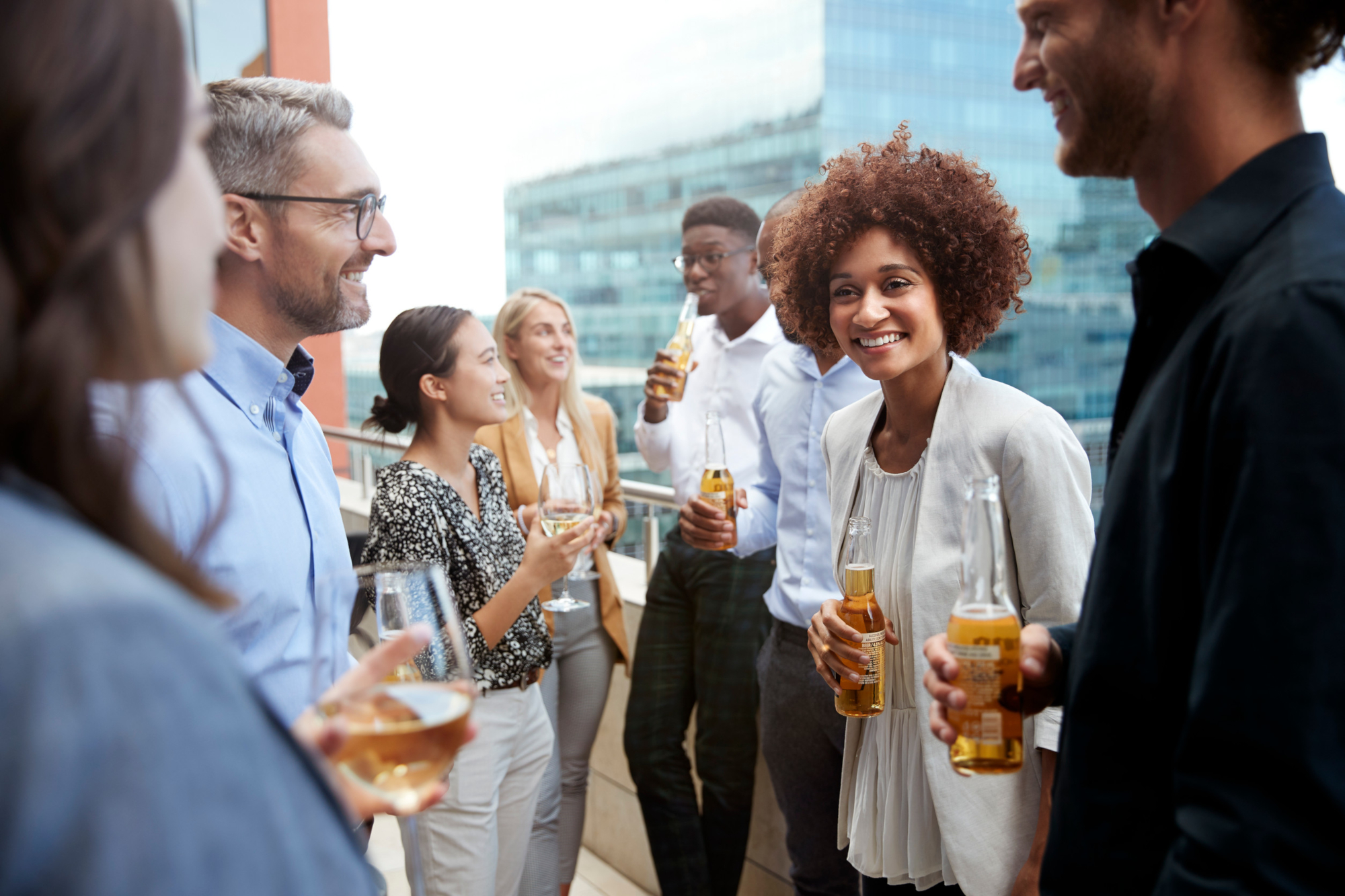 A group of people socializing on a rooftop with drinks in hand, smiling and laughing in front of a cityscape background. Casual, lively atmosphere.