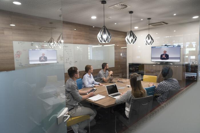 A group of people around a long table, have a video conference with someone on a TV screen